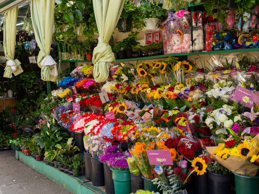A vibrant outdoor floral display featuring an assortment of fresh flower bouquets arranged on a green wooden stand. The arrangement includes sunflowers with large yellow petals and dark centers, white