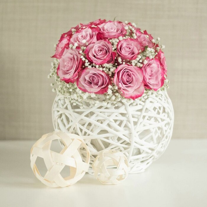 A refined floral still life is captured here, with a rounded bouquet of pink roses and white baby's breath nestled in a decorative white woven vase, flanked by two neutral-toned decorative spheres on a clean white surface. The bouquet itself is generous but compact, with numerous roses gathered closely so that the blooms almost touch, creating a smooth, dome-like silhouette. Each rose displays a delicate gradient of colour: the centres are a soft, almost blush-pink, gradually intensifying to a richer, darker pink at the outer petal edges, giving a subtle two-toned, ombré effect that adds depth without overwhelming the eye. Between the roses, fine sprays of white Gypsophila peek out, their tiny blossoms forming a haze of white that lightens the arrangement and contributes a classic, romantic charm reminiscent of traditional English bouquets often seen at Oxford college celebrations. The vase is a striking, spherical form, crafted from white-painted, interwoven strands arranged in a criss-cross pattern, reminiscent of wicker or rattan yet distinctly contemporary. Its open lattice allows the background colour to show through, creating interesting shadows and adding a tactile, airy feel. Resting on a flawless white tabletop, the arrangement throws a faint reflection, suggesting soft, diffused daylight-perhaps similar to morning light drifting into a quiet townhouse near Headington or along the Iffley Road. To the left, two decorative spheres made of thin, light natural strips echo the woven texture of the vase; one larger, one smaller, they bring a sense of balance and minimalist design. The backdrop is a warm, off-white or beige wall with gentle texture, serving as a neutral canvas that lets the pink roses and white details stand out clearly. Overall, the image conveys calm elegance and understated luxury, perfectly suited to illustrate thoughtful home decor, romantic gifts, or florist-crafted centrepieces in an Oxford setting.