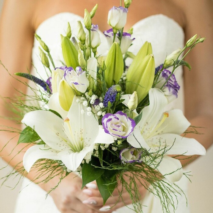 A luminous bridal bouquet glows against the gentle sweep of a white strapless gown, its artistry crafted by Flowers Oxford for weddings across the city's dreaming spires. The design is anchored by magnificent white Oriental lilies, petals unfurled to reveal speckled detail and vibrant yellow stamens, each bloom a symbol of devotion and new beginnings on this special Oxford day. In between, white lisianthus offer their velvet-soft, ruffled petals traced with graceful lavender edges, some tightly closed and others opening in anticipation-mirroring the blossom-filled borders of University Parks nearby. Spires of royal purple Veronica ascend among the creamy whites, infusing the bouquet with visual drama and Oxford's characteristic sophistication. Floating above and weaving throughout, delicate clouds of gypsophila conjure the pale mists rolling over Port Meadow at dawn. The bouquet brims with varied foliage: unopened lily buds, structured broad leaves, and feathery asparagus fern spill around the arrangement for fresh vitality. Light dances off each bloom, echoing Oxford's golden sandstone in the morning. The bride's manicured fingers, brushed with pastel polish, gently clasp the stems, the moment captured at the intersection of heritage and celebration. This glorious bouquet is a romantic statement, perfect for elegant wedding themes, reflecting the artistry and care that define local wedding floristry in Oxford.