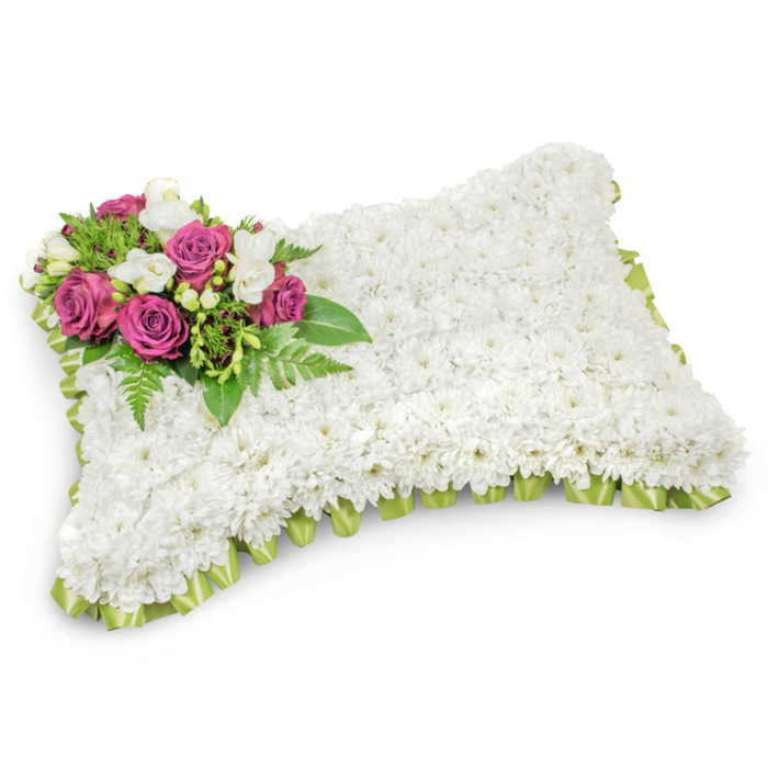 Meticulously assembled for farewell services, this pillow-shaped sympathy arrangement is a study in calm: the entire rectangular form is covered with hundreds of small, immaculate white chrysanthemums so tightly placed that the surface reads like a soft, downy cushion inviting quiet reflection. The pristine white body suggests purity and repose, while a deliberate floral accent sits on the upper-left corner - several deep magenta to fuchsia roses open with velvety layers, symbolising deep affection and remembrance. Interspersed among these roses are sprays of fragrant white freesias and tiny white buds that lift the grouping with both scent and lightness. Beneath and around the focal spray, sprays of glossy green foliage and delicate fern fronds supply texture and a natural frame that softens the transition from the vibrant corner to the cool white expanse. A neat pleated edge of pale green satin ribbon traces the pillow's outline, peeking from under the chrysanthemums and adding a subtle, fresh colour that suggests renewal. Set against a simple white background with a gentle shadow to emphasise its form, the arrangement reads as both traditional and quietly modern. As Flowers Oxford often crafts for services at venues like Magdalen College chapel or mourners gathering near the Covered Market, this cushion-shaped tribute offers a dignified, tender expression of sympathy, conveying peace, rest and enduring memory when families seek a composed floral presence at a funeral or graveside.