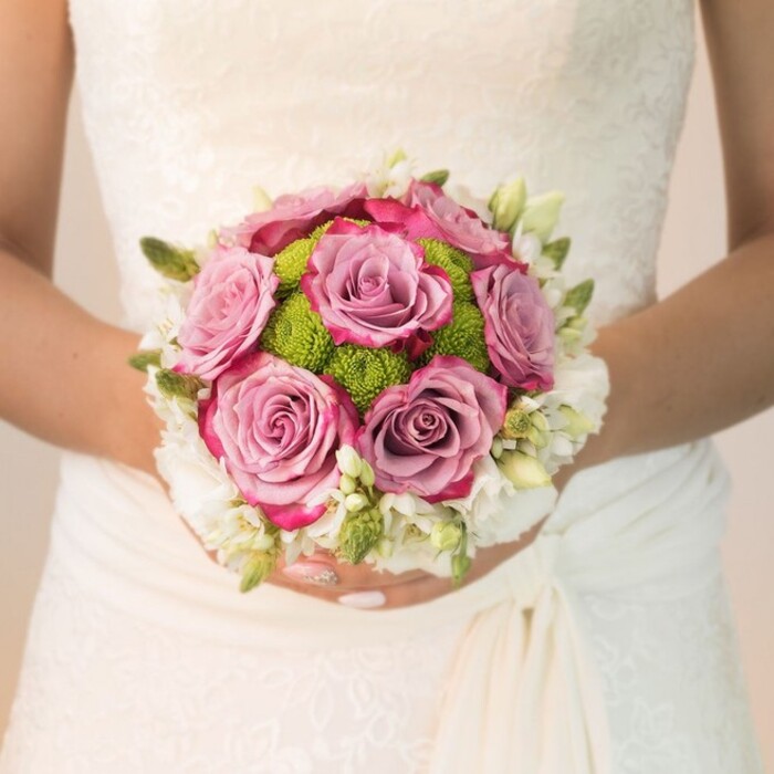 A beautifully detailed, waist-level photograph captures a bride standing in Oxford, holding an elegantly rounded bouquet against her lace-embellished, off-white bridal gown. The bouquet's heart is formed by several grand, full-bloomed roses in hues of dusty mauve to soft lavender, their petals tinged with vibrant magenta and fuchsia along the ruffled outer edges, creating a captivating gradient ideal for Oxford's romantic settings-perhaps near Christchurch Meadow or Port Meadow. Bright lime-green button chrysanthemums appear in tight clusters among the roses, adding vitality and a striking textural play, reminiscent of fresh botanicals sourced from Oxford's renowned floral markets. The bouquet's rim is circled with dainty, pristine white florals-spray rose clusters or delicate freesia-framing the arrangement with gentle purity and complimenting the intricate lacework visible on the bride's sleeves and bodice. Tiny green buds and wisps of leafy texture are artfully interspersed throughout, adding organic depth anchored in traditional English floral design. The bride's hands, gently manicured in a soft pink with a trace of sparkle, cup the bouquet in a pose that speaks of quiet excitement and new beginnings. The gentle, even lighting ensures that every detail-from lace to fresh petal-is captured, embodying the refined craftsmanship, romance, and tradition of Oxford weddings. This lush and intentionally crafted bouquet, as offered by Flowers Oxford, highlights a perfect blend of local inspiration and bridal artistry, designed for those celebrating love amid the spires and gardens of Oxford.