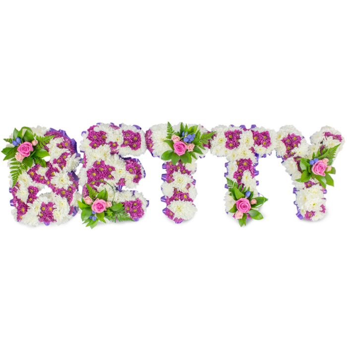 A bespoke sympathy tribute spells the name BETTY in strong, three-dimensional capital letters, each letter densely packed with pristine white chrysanthemum blooms that form a fluffy, textured plane against a pure white studio background. The white chrysanthemums create a calm, almost architectural base; layered within them, vivid magenta to deep purple chrysanthemums trace the inner edges and decorative outlines of each letter, offering contrast and visual rhythm that helps the name read clearly from a distance. Along the outer faces a discreet purple ribbon trims the edges, giving a soft, satin sheen that complements the matte petals. Picking out several focal points, small, natural bouquets are placed strategically - a cluster on the left curve of the B, another at the centre of the first T, and a final accent on the upper arm of the Y - each composed of gentle pink roses in varying openness, tiny bright blue sprayed blooms resembling forget-me-nots or small delphiniums, and lush green foliage such as fern fronds that bring a cooling, leafy texture. The combined palette of white, purple, pink and blue reads as composed and comforting, intended to express deep sympathy while also celebrating a life. The piece is presented with the quiet dignity expected at funerals and memorial services in Oxford, from smaller parish chapels near Jericho to larger services around Magdalen Bridge, and is the kind of personalised floral tribute families select when words feel insufficient.