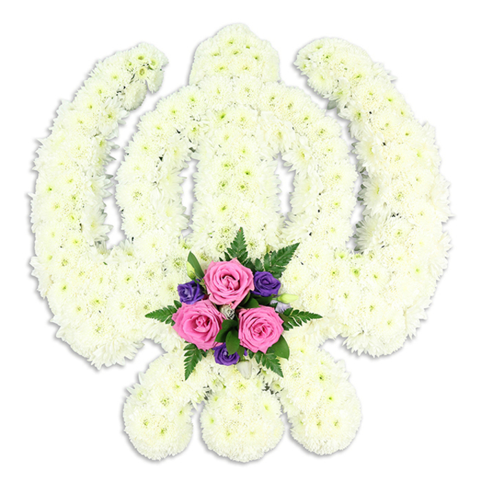 A striking Sikh Khanda floral tribute dominates the frame, its sacred geometry rendered entirely in flowers and set against a bright white background that emphasises every contour. The central double-edged sword rises upright, perfectly balanced within the circular Chakra, while two outward-sweeping Kirpans cross at the base to complete the familiar Sikh emblem. Each part of this complex shape is densely covered in small white chrysanthemum blooms, positioned so closely together that they create an almost sculpted surface. The blossoms are fresh and full, their layered petals forming a soft, cushiony texture that diffuses the light and gives the tribute a serene, almost glowing quality. A faint shadow beneath the design hints at its thickness and three-dimensional structure, as though the Khanda is gently hovering above the background. Nestled precisely where the Kirpans meet at the lower centre is a concentrated burst of colour: three pink roses in full, rounded bloom, their petals spiralling outward, are surrounded by delicate deep purple flowers and finely cut fern foliage in rich green. This accent arrangement appears carefully wired and tucked into place, adding depth and visual focus without overwhelming the simplicity of the white. The contrast suggests love, memory, and gratitude held within a framework of faith. It is the kind of bespoke funeral tribute that might be delivered to a Sikh service in Oxford, perhaps near the city centre or in areas like Cowley and Headington, where families seek meaningful ways to honour their loved one. Created with the precision and care of an experienced local florist, the piece radiates quiet devotion, respect, and enduring spiritual connection.