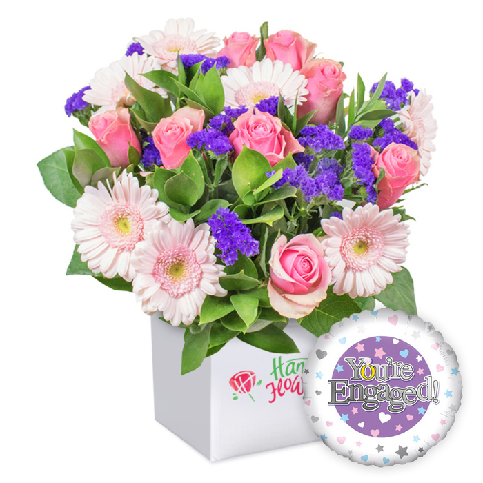 A bright, romantic bouquet from Flowers Oxford stands proudly in a snowy white square box, combining full-bloom pink roses with cheerful light pink gerberas that glow with their soft edges and pale centers. Deep purple statice provides striking pockets of color, while abundant green foliage frames the arrangement, giving it a lush, garden-like vitality. The container's clean, modern lines set off the bouquet's warm palette, drawing the eye to the harmony of pinks, purples, and greens. On the box's front, a subtle red rose icon paired with green script suggests a local, trusted florist's mark without naming the brand. To the lower right, a round foil engagement balloon floats, its surface dominated by a central purple circle, silver lettering declaring You're Engaged! and a white ring rimmed with pastel hearts and silver stars, with a gleaming gold 'O' accenting the text. This elegant, contemporary gift is ideal for celebrating an engagement in Oxford's Jericho, North Oxford, or university-adjacent communities, translating joy into a lasting bouquet. It's designed for reliable, timely delivery across the city, brightening homes from Oxford's historic High Street to the leafy suburbs of Headington with a confident, warmth-filled message of love and new beginnings.