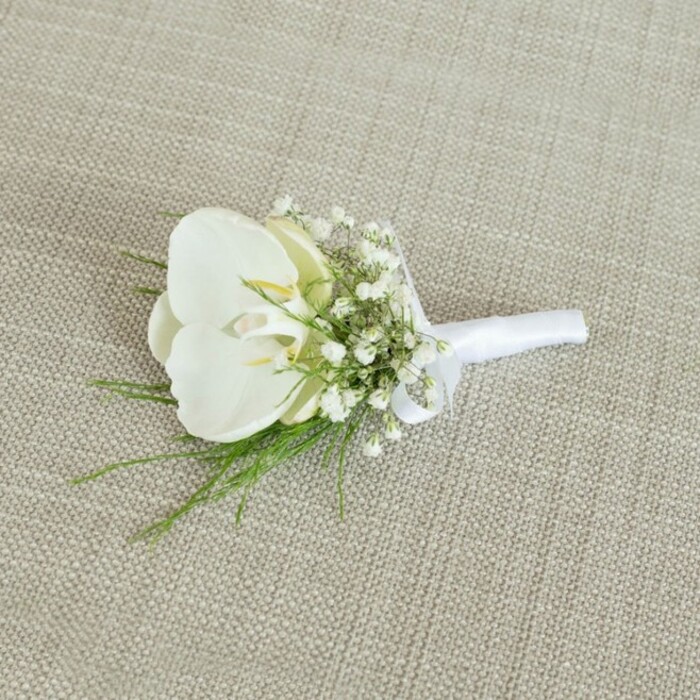 A sophisticated white orchid boutonniere is shown resting on a pale beige, linen-like fabric, the subtle woven threads beneath it adding a gentle, tactile backdrop that enhances the arrangement's delicate details. At the centre, a single Phalaenopsis orchid bloom unfurls in full, relaxed openness, its petals a cool, creamy white with a soft, velvety sheen, like pressed silk. Nestled in the throat of the orchid, a faint wash of pale yellow and cream draws the eye inward, hinting at the flower's natural structure and vitality. Encircling this central bloom, fine sprays of white baby's breath create a misty, cloud-soft border, each minuscule flower contributing to a sense of lightness and romantic air. Slender, arching tendrils of fresh green foliage weave through and just beyond the gypsophila, the bright green tones suggesting new growth along the riverside paths that curve through Oxford. The stems are brought together with the precision of an experienced florist, then wrapped tightly in a smooth, matte white satin ribbon that covers the binding and leads to a small, understated bow, carefully positioned to sit flat when pinned to a jacket lapel. The overall lighting is calm and evenly diffused, bringing out the crisp contrast between the white petals, the green foliage, and the warm, neutral background without glare or harsh shadow. The composition suggests a timeless wedding accessory, perfect for a groom, best man, or guest at an Oxford ceremony in a college chapel or registry office, where subtle fragrance, clean lines, and thoughtful craftsmanship come together in a miniature expression of elegance and new beginnings.