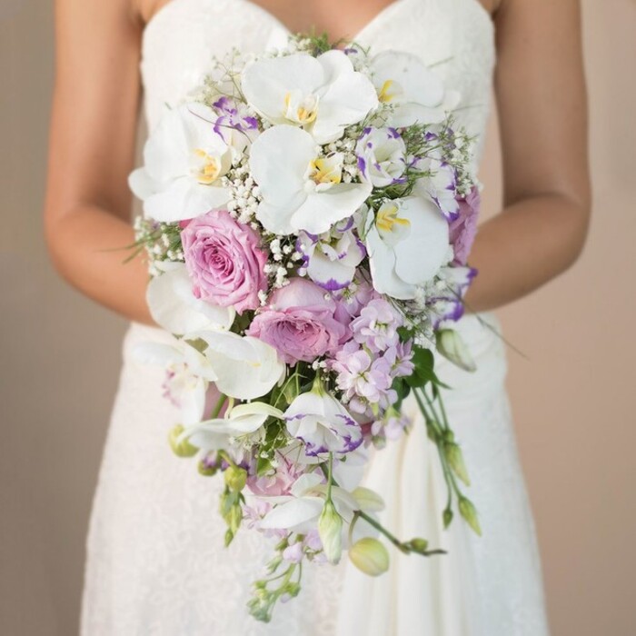A captivating bridal bouquet, crafted to perfection by a seasoned Oxford florist, fills this vertical photograph with a sense of romance and artistry. Set against a tranquil, blurred beige background that echoes the parchment tones of Oxford's ancient libraries, the bride is shown from the chest down, entwined gently around a dramatic, flowing bouquet. Her classic strapless gown features delicate white lacework, its intricate patterns catching the softest morning light found on Magdalen College grounds. The bouquet's silhouette descends gracefully, following a sophisticated teardrop shape that suggests movement and vitality-like a river streaming beneath the city's iconic bridges. Leading the visual ensemble are several immaculate white phalaenopsis orchids, their luminous petals curling outward to reveal golden-yellow and blush centers. Nestled among these are generous, tightly swirled pink garden roses, their velvet texture contrasting beautifully with the nearby points of lavender stock and white lisianthus edged in royal purple. Sprays of baby's breath gently fill the negative spaces, creating an impression of airiness and volume, while unobtrusive green leaves peek through, adding just a hint of woodland charm reminiscent of Oxford's leafy streets. Trailing tendrils and unopened orchid buds extend below, giving the entire bouquet a sense of organic flow and fresh vitality. This bridal arrangement, ideal for an elegant Oxford ceremony, reflects the region's commitment to tradition and artisan skill, making it a cherished highlight for couples celebrating amidst the city's storied spires.