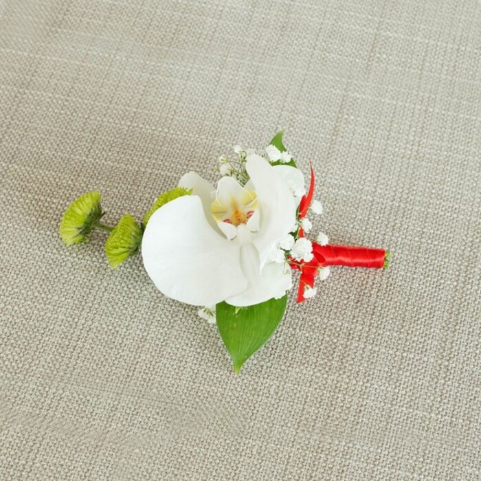 An elegant boutonniere is displayed against a softly textured, light beige linen backdrop, the fabric's fine yet visible weave evoking the natural, understated charm often found in Oxford's historic venues and converted barns along the edge of town. At the centre of this small composition is a pristine white phalaenopsis orchid, its large petals unfurling with a gentle, sculptural grace. The surface of each petal looks almost satin-smooth, reflecting the light in a subtle, diffused glow. Within the orchid's throat, delicate touches of soft yellow blend into hints of reddish-orange or light brown patterning, fine markings that add warmth and a sense of intricacy to the bloom's otherwise minimalist palette. Surrounding the orchid are loose, cloud-like clusters of baby's breath, their tiny white flowers creating an ethereal halo and softening the line between blossom and background. To the left, two small, spherical lime-green chrysanthemum buds offer a striking contrast, their densely packed, spiky petals resembling tiny pom-poms of fresh foliage, crisp and vibrant. Beneath the flowers, a broad, deep green leaf curves gently, forming a natural base that grounds the design, while a smaller, darker leaf emerges near the stem, adding layered green tones like those found in shaded borders of Oxford's Botanic Garden. The stems of the arrangement are tightly wrapped in a rich crimson red satin ribbon, creating a smooth, polished handle. The ribbon is finished with a neat knot or discreet bow just under the flower heads, introducing a bold, celebratory dash of colour that stands out against both the neutral fabric and the white petals. Crafted with the attention to detail you would expect from an experienced local florist, this boutonniere feels tailored for a groom or groomsman at an Oxford wedding, combining classic orchid elegance with contemporary touches suitable for black-tie events, civil ceremonies, or intimate celebrations in the city's charming hotels and colleges.