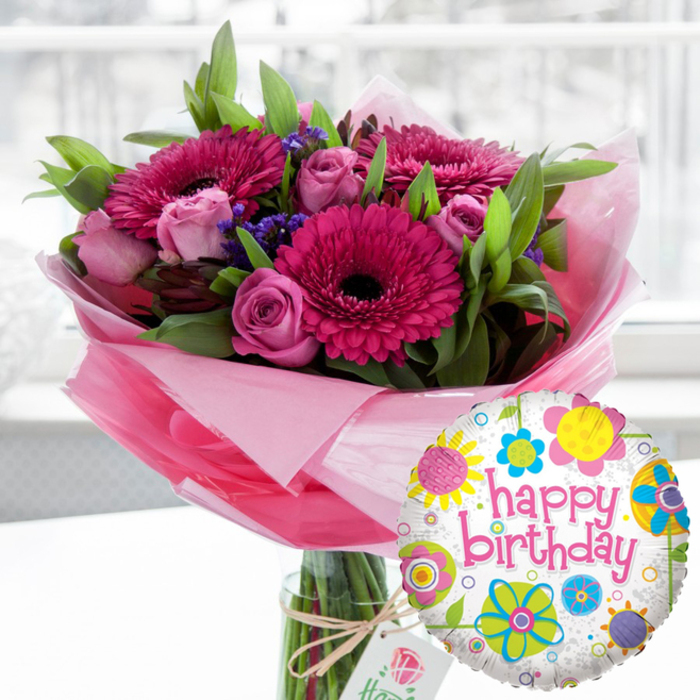 A joyful birthday scene is captured in soft focus, highlighting a lavish pink bouquet and a cheerful foil balloon that could easily be sitting on a dining table in an Oxford terrace house near Jericho. The bouquet is arranged in a tall, clear glass cylinder vase, the green stems and water visible through the sides, emphasising how freshly the flowers have been gathered. Around the base of the blooms, translucent light pink cellophane is folded and flared, creating a gently ruffled collar that softens the line between flowers and glass. Dominating the display are large gerbera daisies in rich tones of fuchsia and magenta, their petals perfectly layered around deep, almost black centres that add dramatic contrast. Threaded between the gerberas are numerous roses in shades of blush and mid-pink, some still tight and rounded like little promises, others beginning to open and reveal their spiralled hearts. Small tufts of deep purple filler flowers, likely statice, peek out between the larger blooms, bringing a cool note to the vivid palette. Lush, bright green foliage, with both broad and slender leaves, creates a sense of fullness and movement, giving the arrangement a natural, garden-picked feel even though it is clearly crafted with expert care. A length of rustic twine is tied around the vase's neck, securing a small, off-white tag stamped with a delicate rose emblem, the sort of detail a local florist like Flowers Oxford would choose to complement a premium birthday gift. Just in front and slightly to the right, a round Mylar Happy Birthday balloon completes the ensemble. Its white background, flecked with pale grey, is scattered with playful stylised flowers and circular motifs in sunny yellow, bright blue, lime green, vivid pink, and deep purple. Across the middle, in bold pink cursive, the words happy birthday curve happily, mirroring the tones of the gerberas and roses. The softly blurred indoor setting and natural light create an inviting atmosphere that focuses attention on this uplifting birthday arrangement, ideal for surprise deliveries across Oxford.