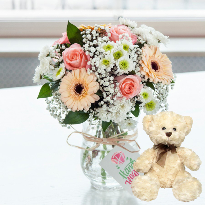 A softly glowing anniversary arrangement is set on a pristine white surface, reminiscent of a bright kitchen table in an Oxford townhouse near Summertown. In the centre stands a clear glass jar-style vase, its rounded shape and visible stems giving a relaxed, homely feel. The vase cradles a generous bouquet of pastel flowers: blush-pink roses with gently unfurling petals, peach-toned gerbera daisies accented by deep brown centres, and clusters of small white chrysanthemums whose lime-green cores add a fresh pop of colour. Wisps of white baby's breath thread between the larger blossoms, lending an airy lightness that feels like early spring along the River Cherwell. Around the neck of the vase, a band of natural twine is tied in a simple bow, from which dangles a neat rectangular gift tag. The tag is white with a pink heart or gemstone-like emblem and soft green script suggesting a handmade or personalised sentiment, as if a local florist had penned a message before a same-day delivery. To the right of the bouquet, a cream-coloured teddy bear sits upright, angled slightly toward the flowers. Its plush fur is textured and inviting, its dark bead eyes and brown nose adding a sweet, almost shy expression. A slim, dark brown satin ribbon is tied neatly around its neck, echoing the warmth of the gerbera centres. Behind them, the background is softly blurred, implying a bright window and letting the composition remain calm and uncluttered. Together, the fresh bouquet and teddy bear form a tender anniversary tableau, ideal for surprising someone in Oxford, from Cowley Road flats to quiet homes in Botley, with a gesture of enduring affection.