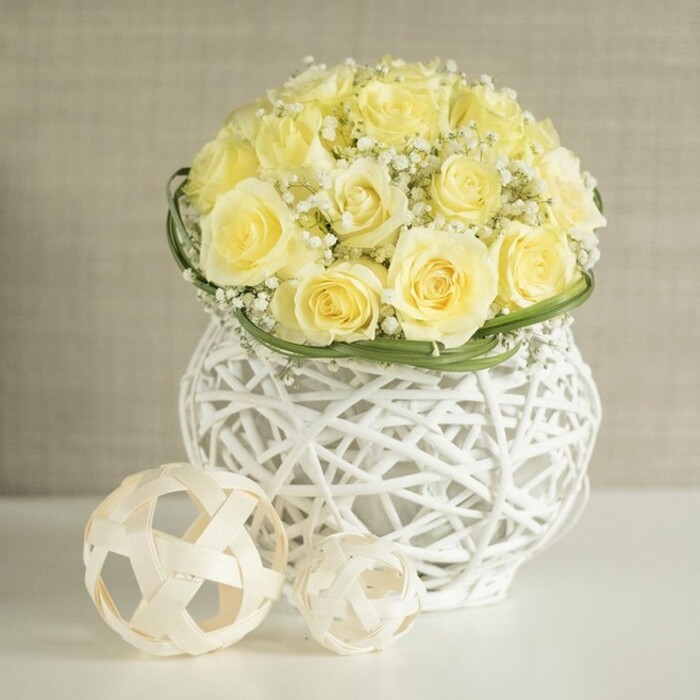 On a smooth white table, a sophisticated wedding table arrangement draws the eye: a lush, dome-shaped bouquet of pale yellow roses, each blossom carefully turned outward so the full swirl of petals can be admired. The roses are so closely nestled that they form a continuous, velvety surface, their buttery tones softened by sprays of white baby's breath threaded throughout like tiny stars in a spring sky. Just below the blooms, a band of vivid green leaves has been woven neatly around the base, creating a tidy collar that hints at careful craftsmanship and adds a fresh, natural outline. The bouquet is presented in a striking white woven vase, its open lattice reminiscent of contemporary wicker or rattan, with curved lines that give it an organic, almost sculptural presence. Light filters through the gaps in the weave, casting subtle shadows onto the table and echoing the gentle interplay of sunshine and shade you might see in an Oxford courtyard garden off Iffley Road or in leafy North Oxford. Beside the vase rest two pale, intertwined wooden spheres of different sizes, their openwork design mirroring the texture of the vessel and adding a modern decorative touch without competing with the flowers. The background remains softly blurred in warm neutral tones, calming and understated, so the delicate yellows, crisp whites, and fresh greens remain the star of the scene. The overall mood is one of graceful celebration and quiet joy, ideal for dressing wedding breakfast tables in Oxford hotels, intimate receptions near the Covered Market, or elegant anniversary dinners at home with flowers delivered freshly arranged by a trusted local florist.