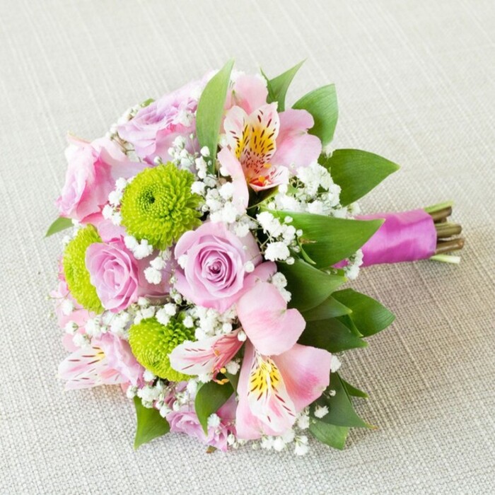 A lush, hand-tied bouquet commands attention as it rests diagonally on an understated, light beige backdrop reminiscent of Oxford's historic stone facades. Soft pastel tones of blush-pink and lilac roses form the focal point, each blossom's spiraled petals quietly suggesting both beauty and longevity-a mirror to Oxford's enduring elegance. Interspersed among these are cheerful lime-green button chrysanthemums, their compact, textured globes adding a bold twist of colour and tactile interest reminiscent of the city's spring gardens. Accentuating the design are pale pink alstroemeria (Peruvian lilies), each flower opened wide to display subtle yellow and deep burgundy markings, infusing the bouquet with a hint of wild charm. Between these larger blooms, the bouquet is softened by cloudlike clusters of white gypsophila, while deep, glossy green leaves nestle throughout, enhancing both structure and freshness. Binding the natural green and brown stems is a lustrous, magenta-purple satin ribbon, wrapped securely and left with tidy tails-a finish suited to modern weddings or stylish anniversary celebrations along Oxford's High Street. The overall composition radiates happiness, sophistication, and the romantic spirit found in local celebrations. Available for same-day delivery by Flowers Oxford, this bouquet offers a touch of Oxford's timeless beauty, perfect for brightening any home from Jericho to Cowley with its fragrant, artistic presence.