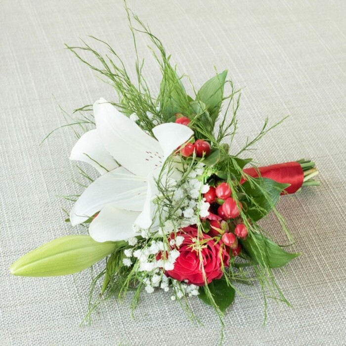 In this refined Oxford-inspired bridesmaid bouquet, an open white lily commands attention with its serene petals stretching outward and warm yellow anthers tucked deep within, the center kissed with delicate flecks of rusty brown. Resting upon the textured, light beige linen typical of Oxfordshire's gracious interiors, the bouquet's central bloom is accompanied by a lavish, full red rose-its dense, velveteen spirals exuding heartfelt celebration much like a festive gathering at the Sheldonian Theatre. Sprigs of airy white gypsophila (baby's breath) intermingle among the major flowers, casting a gentle, cloudlike veil, while clusters of glossy red berries intersperse the arrangement, adding cheerful vibrance reminiscent of the winter berries found along Addison's Walk. A verdant lily bud unfurls quietly at the edge, hinting at future blossoms and stories yet to be written. Surrounding this ensemble, an assortment of green foliage creates depth-some fronds fine and fern-like, others broad and emerald-crisp, conjuring memories of riverside walks along the Cherwell. The bouquet's base is elegantly finished with a deep crimson satin ribbon, its sheen harmonizing with the red berries and rose while imparting an uplifting sense of festivity. Soft, diffused daylight caresses the arrangement, bringing every petal and leaf into gentle focus. Meticulously crafted for momentous occasions, this bouquet resonates with the tradition and warmth unique to celebrations in and around Oxford, suitable for weddings, anniversaries, or as a cherished, eye-catching gift.