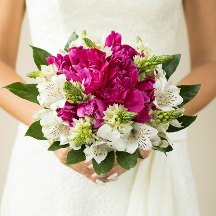 In this detailed bridal close-up, a bride stands in her white lace wedding dress, holding a rounded bouquet that feels both modern and timeless, the kind of design often requested for Oxford weddings in Jericho churches or marquee receptions near Port Meadow. Central to the arrangement are generous peonies in shades of intense fuchsia and magenta, their full, ruffled petals forming a dense, velvety cluster that immediately draws the eye. Each peony head appears at different stages of opening, some tightly cupped and others fully unfurled, creating depth and natural movement. Interwoven throughout are crisp white alstroemeria, or Peruvian lilies, their slender petals flaring outward and decorated with delicate maroon speckling and fine markings that lend an intricate, almost lace-like pattern to the bouquet, echoing the gown's fabric. Slim, spiky green buds and textured filler flowers stand upright among the softer blooms, introducing a playful contrast in shape and a fresh, just-picked energy reminiscent of early summer mornings in the Oxford Covered Market flower stalls. Encircling the flowers, rich dark green leaves provide a structured frame, their broad surfaces and subtle sheen grounding the vibrant colours and ensuring the bouquet keeps its neat, rounded profile. The bride's hands are visible at the base, gently clasping the stems, with light pink polished nails that subtly mirror the warmth of the peonies. The background is softly blurred and light, ensuring the vivid hues and careful craftsmanship remain the focus. Overall, the bouquet radiates joy, confidence, and romance - a striking choice for a spring or summer ceremony anywhere in Oxford, from historic college quadrangles to intimate garden venues. Created in this style, it celebrates both bold colour and delicate detailing in perfect balance.