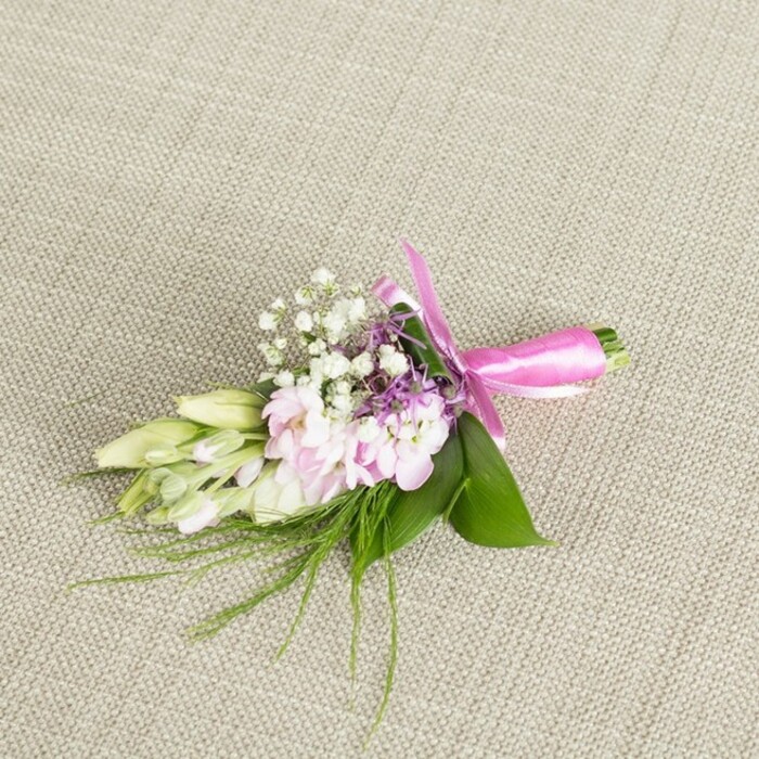 A petite yet striking floral boutonniere is photographed against a softly woven, neutral fabric, the colour somewhere between light oatmeal and warm stone, much like the honeyed walls of central Oxford. Centered on this calm backdrop, a cluster of blush pink flowers forms the heart of the piece, their many small, ruffled petals layered closely together to mimic miniature stocks or garden-style blooms. The pink shades are gentle, moving from pale, almost shell pink at the outer petals to a slightly deeper tone inside, creating a subtle depth without overwhelming the eye. Dotted through the pink cluster are sprigs of white baby's breath, their tiny cup-shaped flowers on thin green stems adding a delicate spray of texture, as if fine lace has been tucked into the arrangement. Around and between these, a few small spikes of lavender or deep purple florets provide a cooler contrast, hinting at English cottage gardens and borders seen along the paths by the River Cherwell. On one side, creamy white to pale green buds, likely freesia or a similar narrow-petalled flower, lie closed but plump, Nods to freshness and anticipation-as if they will open just in time for an afternoon ceremony at a college chapel. The base of the arrangement is anchored by broad, glossy green leaves that fan gently outward, while fine, grass-like greenery and wispy foliage extend in soft arcs, giving a light, natural movement. The stems are snugly aligned and wrapped in a vivid fuchsia satin ribbon, the fabric smooth and reflective, knotted into a neat bow with gracefully curving loops. The overall feel is of a carefully hand-tied buttonhole, perfect for weddings, proms, or black-tie events across Oxford's historic venues, crafted with an eye for both elegance and modern colour harmony.