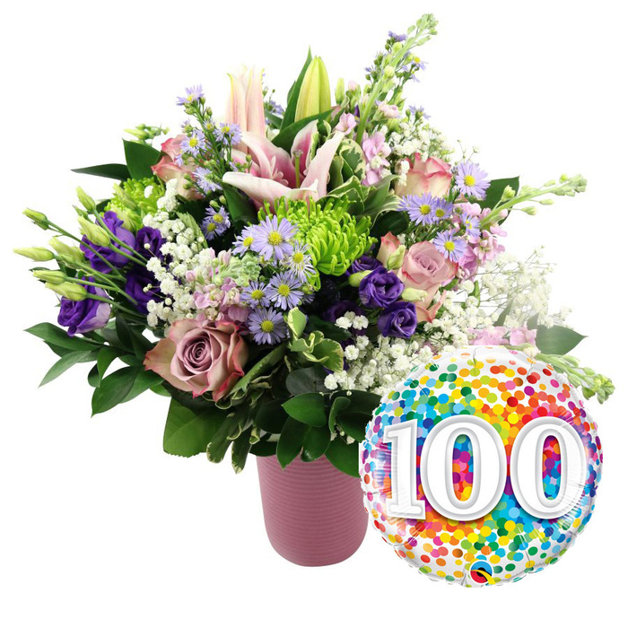 A radiant centenary celebration scene unfolds against a spotless white background, where a generous floral bouquet in a stylish pink vase is partnered with a colourful 100 balloon. The vase itself is a soft dusty-rose shade with a ribbed, matte texture, lending a modern yet gentle feel that frames the flowers without overpowering them. Rising proudly from the centre are pale pink Oriental lilies, some fully open into starburst blooms, their petals curling slightly at the tips, others still nestled in bud form, suggesting continuity and life still unfolding even after a hundred remarkable years. Around these focal blooms, a mix of light pink roses and richer mauve or dusty-rose roses form a lush, fragrant collar, their velvety petals layered in soft spirals. Delicate purple lisianthus, with their ruffled edges, and clusters of small lavender asters with bright yellow centres provide spirited touches of colour that recall springtime borders in Oxford's Botanic Garden. Bright lime green button chrysanthemums punctuate the arrangement like little fireworks, while airy sprays of white baby's breath drift between stems, giving the bouquet a cloud-like softness. Varied deep green foliage, from broader leaves to slender stems, supports and frames the blossoms, creating a sense of fullness and natural movement as if it were just arranged in a local Oxford florist's studio. To the lower right, a round foil balloon reflects the light, completely covered in rainbow confetti dots of red, orange, yellow, green, blue, and purple. Bold white numerals 100, slightly shadowed for depth, stand proud at its centre. The composition together speaks of gratitude, longevity, and pride, ideally suited for marking a 100th birthday, a century-old institution, or a special Oxford family milestone that spans generations.