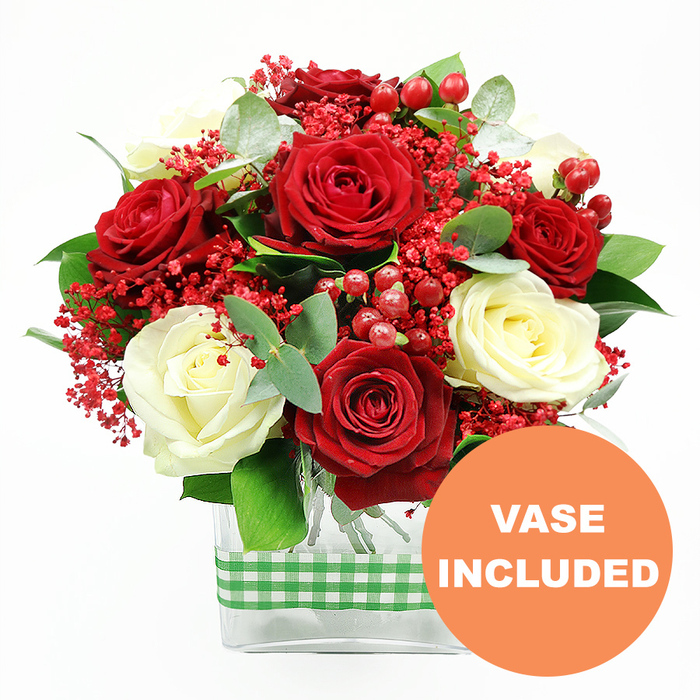 This image presents a carefully styled, celebratory bouquet of roses in red and white, photographed against a spotless white background that allows each colour and texture to stand out. Dominating the composition are velvety, fully opened red roses, their rich colour and layered petals suggesting romance and warmth. Interwoven among them are creamy white roses, slightly softer in tone, balancing the bolder red with a sense of calm and grace. Small clusters of vivid red filler blooms, similar to dyed gypsophila, scatter throughout the arrangement like tiny confetti, lending an airy, almost cloud-like texture above the more substantial blossoms. Nestled between the petals, glossy red hypericum berries offer a sculptural, bead-like contrast, catching light and creating points of shimmering interest. Surrounding the blooms, a mix of lush green foliage and cool eucalyptus leaves creates a natural frame, the varied shapes and subtle bluish tones of the eucalyptus adding depth and a modern twist. All stems are neatly arranged in a clear rectangular glass vase, whose transparent sides reveal the vertical lines of the gathered stems, underscoring the clean, contemporary style many families in Oxford enjoy in townhouses from Summertown to Iffley. Around the middle of the vase, a green and white gingham ribbon wraps smoothly, suggesting rustic comfort and seasonal charm, suitable for Christmas dinners, winter birthdays, or a summer anniversary supper at home. In the bottom right of the image, an orange circular emblem with the words VASE INCLUDED in bold white letters confirms that this floral design is supplied as a complete, ready-to-display piece. The overall mood is one of joyful sophistication, ideal as a romantic gesture or a thoughtful gift for loved ones across the Oxford area.