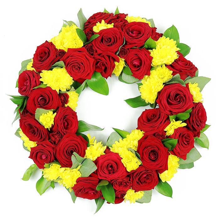 Against a stark white background, a lush circular wreath for a funeral or memorial service is shown in full, its flowers arranged with evident professional care. The wreath forms a complete, unbroken ring, densely packed with deep red roses and bright yellow chrysanthemums that sit closely together, creating a rich tapestry of colour and texture. The roses dominate the composition, their petals thick and velvety, unfolding from tightly coiled centres into layered cups that vary in shade from classic ruby to a more intense burgundy, lending the arrangement depth and shadow. Nestled among them, sunny yellow chrysanthemums cluster in small groups, each head made up of many delicate, thread-like petals that give a soft, almost fluffy appearance. These yellow blooms lighten the mood of the piece, suggesting remembrance not only tinged with sorrow but also with gratitude and celebration of a life. Beneath and around the flowers, an array of greenery forms a subtle frame: broad, rounded leaves with a glossy finish likely come from salal or ruscus, while cooler, more matte sprigs resemble eucalyptus, their silvery tone adding a calming note. The foliage is carefully placed so that it creates a natural border without disturbing the wreath's perfect circle. The lighting is bright and neutral, making the reds, yellows, and greens appear vivid but not harsh, as if the wreath has just been completed in a local Oxford florist's workshop near Summertown or Jericho, ready for delivery to a church, crematorium, or family home. The overall feeling is one of dignity and tenderness, a floral tribute that speaks of enduring love, heartfelt sympathy, and the wish to honour someone's memory with beauty that feels both classic and personal.
