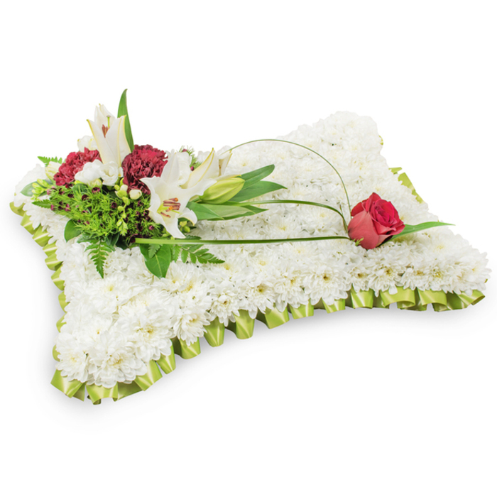 A reverent, pillow-shaped funeral tribute made entirely from tightly packed white chrysanthemums rests against a neutral white background, its rectangular silhouette conveying comfort and repose. The entire perimeter is trimmed with a refined pleated ribbon in a delicate lime green satin, the pleats forming a soft frill that gives the piece a finished, respectful appearance. On the upper left, a composed cluster of accent blooms provides the focal point: two open white Oriental lilies with their characteristic central stamens and a graceful closed bud, paired with dark burgundy spray carnations whose textured, ruffled petals introduce a deep, consoling tone. The cluster is set against an abundance of green foliage - airy fern fronds and broad salal leaves - which lends structure and the scent of fresh greenery. From this grouping, a narrow arch of bright green bear grass or similar leaf arcs thoughtfully across the cushion, leading the gaze to a single deep red rosebud poised on the upper right edge, its velvety form just beginning to unfold. The juxtaposition of pure white blooms, somber red accents and soft green trim creates a palette of purity, love and remembrance. The lilies give a faint, sweet perfume while the chrysanthemums offer a plush, tactile surface evocative of a soft pillow. This traditional sympathy design is suitable for memorial services and funeral settings in Oxford - whether being delivered to college chapels on High Street or to services near the Thames (the Isis) - and expresses deep condolence with measured elegance and experienced florist care.