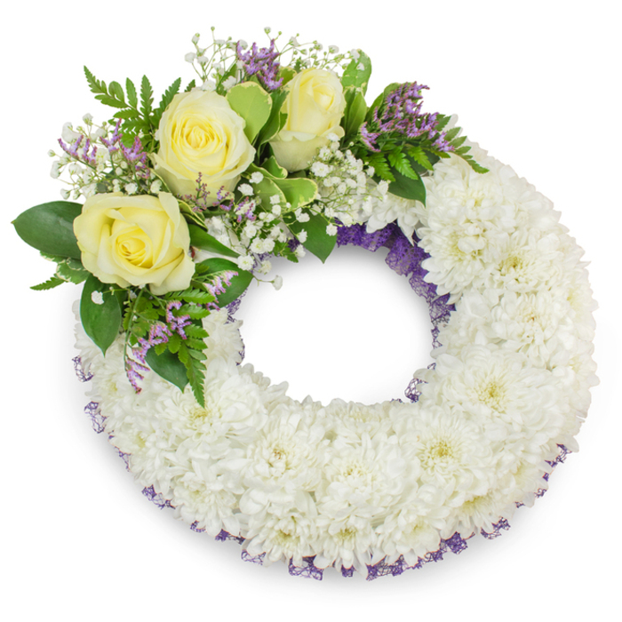 A dignified circular wreath formed from hundreds of small, immaculate white chrysanthemums creates a calm, continuous ring that reads as a tactile symbol of peace and ongoing remembrance. The chrysanthemum heads are packed densely so the surface appears plush and inviting, their cool white petals lending a soft, quiet clarity under the photograph's flat, neutral lighting. Narrow bands of deep purple ribbon or fine mesh are intentionally visible along the inner and outer edges, their colour offering a discreet but sophisticated frame that deepens the wreath's visual weight. Anchoring the design in the upper-left is a cluster of three creamy yellow roses - petals layered with velvety richness - surrounded by airy stems of gypsophila and small sprays of lavender-hued limonium or statice, which introduce a refined pop of purple. Intervening greenery, from broad glossy leaves to delicate fern-like foliage, provides texture and a verdant backdrop that makes the lighter blooms sing. The composition keeps a respectful balance between formality and gentle warmth; it smells faintly of rose and fresh green leaves, and its proportions are suitable for a funeral or memorial setting. Crafted with the care of an Oxford florist familiar with local needs - Flowers Oxford prepares tributes intended for simple, heartfelt moments of farewell, whether for services near the Covered Market or a quiet ceremony by University Parks - this wreath conveys solace, honour and a peaceful, enduring presence.