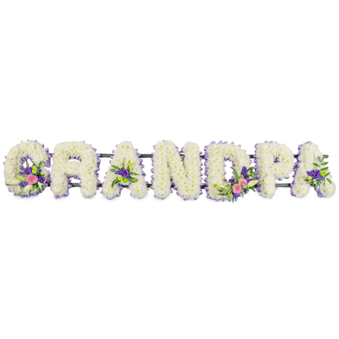 This photograph features a detailed floral name tribute spelling out GRANDPA, a classic funeral arrangement often chosen by families in Oxford to honour a cherished grandfather. The word is presented in a clean horizontal line, with each of the seven capital letters shaped from an abundance of small, pristine white chrysanthemums. Their tightly clustered, multi-petalled heads create a plush, almost quilted surface, giving the letters a soft, compassionate presence rather than a stark, graphic look. Tracing the edge of every letter is a narrow band of petite lilac and purple flowers, probably statice or similar filler blooms, which add fine definition and a gentle wash of cool colour, like a lavender frame around each snowy white form. Several letters carry added focal sprays that bring movement and individuality: the G shows a delicate white lily-like flower rising from a nest of bright pink and fuchsia blossoms with hints of deep purple and glossy green foliage; the first A and the final A each display more modest yet still vibrant groupings of purple and pink flowers; and the letter D boasts a slightly fuller cluster echoing the G, balancing the composition visually from left to right. All the letters are anchored to a slim, dark green bar that runs unobtrusively along the back, keeping them aligned for display on a hearse, in a church, or at a graveside. Set against a seamless white background with soft, diffused lighting, every nuance of texture and hue is visible, creating an impression of calm respect and personal devotion, the sort of tribute you might order locally to accompany a service at Oxford Crematorium or a quiet burial in one of the city's historic churchyards.