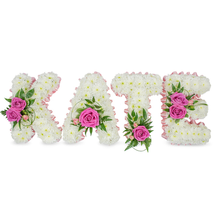 This personalised floral display presents the name KATE in bold, three-dimensional letters, each letter lovingly composed of hundreds of small, pure white chrysanthemums to create a seamless, pillowy surface. The inner edges of every letter are traced with a pale pink satin ribbon that adds an understated, elegant outline and a soft sheen against the white blooms. Carefully positioned clusters of hot pink roses punctuate the otherwise pristine surface: on the K two opened roses sit to the left, the A features a single statement rose near its base, the T carries one bright rose at its centre-bottom, and the E holds two roses along its right-hand curve. Around these focal roses sit delicate pale-pink sprigs-tiny buds and perhaps alstroemeria-together with varied greenery: crisp variegated leaves, fern-like fronds, and glossy ruscus or eucalyptus that lift the composition and introduce cool, verdant tones. The letters are freestanding and cast gentle shadows on the white background, lending depth and a sculptural presence appropriate for placement beside a memorial photograph or chapel lectern. Scent is softly present: the clean, powdery aroma of chrysanthemums underpinned by the sweeter, warmer perfume of the roses. The crafted balance of white, pink and green gives a mood of tender respect and quiet affection. Seen in the calm morning light of an Oxford memorial-perhaps at a service near High Street or a small remembrance in Headington Hill-the piece reads as a heartfelt tribute, a clear and personalised expression of love and remembrance, executed with careful florist technique and compositional restraint.