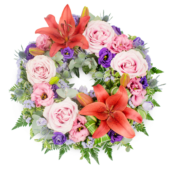 This photograph shows a meticulously crafted circular funeral wreath set against a pure white background, allowing every detail of the design to stand out clearly. The wreath feels both lush and carefully controlled, like a piece created for a thoughtful farewell at a memorial in central Oxford or a quiet gathering near the Botanic Garden. Two large coral-orange to reddish-orange Asiatic lilies serve as the main focal points, one displayed toward the upper arc of the circle and another positioned lower down. Their star-shaped blooms open wide, revealing intricate stamens at the centre, their petals gently curved and lightly textured, radiating a warm, dignified energy.   

Between and around these lilies, an abundance of roses in soft blush and lavender-pink shades fills the ring, each flower fully open, with gently spiralling petals that suggest tenderness and grace. Intermixed are eustoma (lisianthus) flowers in two main tones: a delicate, almost pastel pink-purple and a rich, velvety deep purple. Their ruffled edges create subtle shadows and depth, echoing the layered history of the city's old stone colleges. Scattered throughout are tiny clusters of lavender-blue filler flowers, possibly statice, which add a light, airy haze of colour that softens the overall appearance.   

Spiky blue-grey eryngium thistles punctuate the softer blooms, their bristled heads and slender stems contributing a cool, structural contrast. The floral elements rest on a deeply textured bed of foliage: silvery eucalyptus leaves, slender fern fronds, and dark glossy greens overlap to create a full, verdant frame. Several unopened lily buds peek from within the greenery, suggesting the gentle unfolding of future days. Near the lower lily, bright green leaves are folded and twisted into elegant shapes, highlighting the artisan skill behind the design. This wreath, ideal for same-day condolence delivery across Oxford, visually expresses remembrance, respect, and the unbroken circle of love.