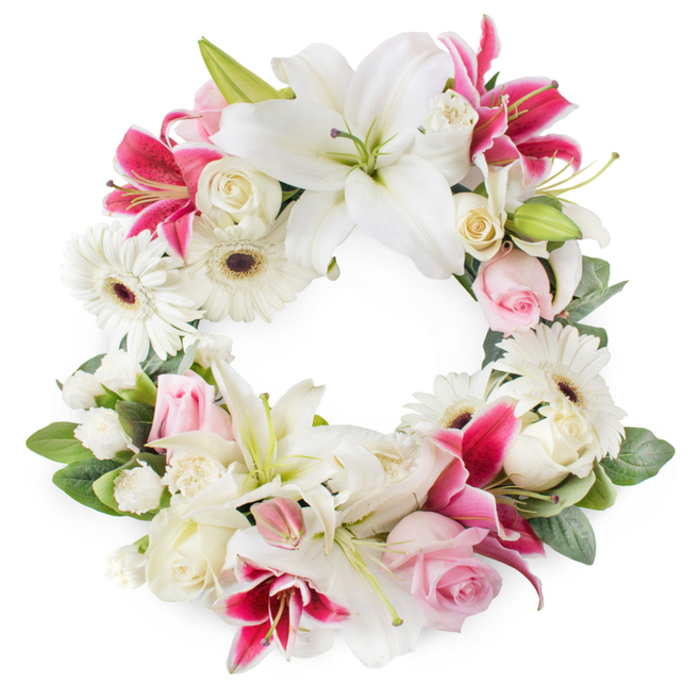 A beautifully composed circular funeral wreath is presented against a bright white background, allowing every bloom and leaf to stand out clearly. The wreath is densely adorned with large white lilies, their elegant petals opening wide and slightly ruffled at the edges, each bloom encircling prominent golden stamens and cool green pistils that lend a sense of purity and light. Among these serene lilies sit vivid deep pink and fuchsia lilies, with star-shaped flowers and softly speckled throats that provide rich, jewel-like accents throughout the ring. Interspersed between the lilies are crisp white gerbera daisies, their layered petals radiating from dark chocolate or reddish centres, giving the design a contemporary yet gentle feel, much like the blend of old and new you find walking through Oxford's historic streets and modern quarters. Nestled in clusters are soft pink roses and creamy white roses, some tightly furled, others fully open with intricate whorls of petals, creating a sense of unfolding remembrance and affection. Smaller white filler blooms, possibly miniature carnations or spray chrysanthemums, are tucked carefully into the spaces, creating a plush, cushiony texture that makes the wreath feel generous and complete. Encasing the florals is a halo of lush green foliage, with varied leaf shapes adding movement and subtle shadow, reinforcing the circular symbolism of eternity and ongoing love. This thoughtfully balanced sympathy wreath, reminiscent of tributes chosen for services near Oxford's leafy parks or quiet chapels, conveys grace, compassion, and a gentle, dignified farewell for a loved one.