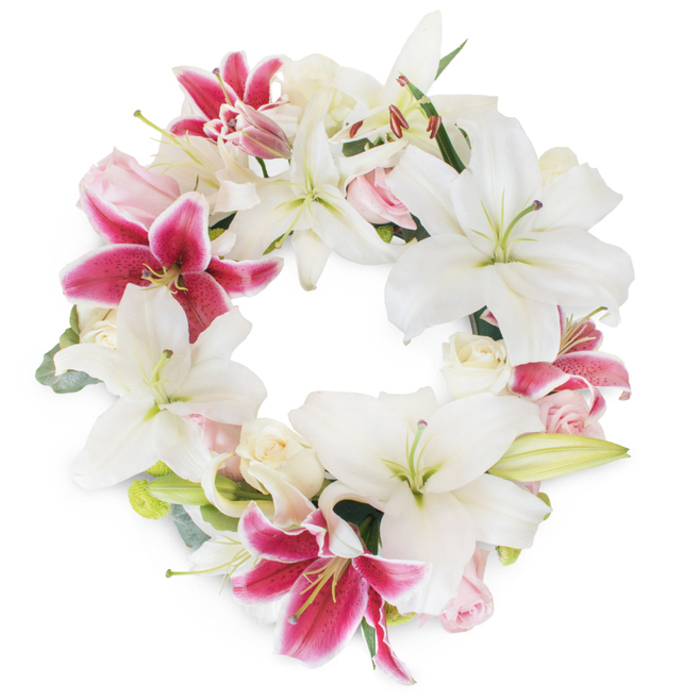A radiant and dignified floral wreath, artfully arranged by Flowers Oxford's skilled florists, commands attention with its lush display of pristine white lilies and dramatic fuchsia tones. The circle is thick with full, star-shaped white lilies whose gently curved petals and prominent stamens create a foundation of purity and peace-perfectly suiting memorials held in tranquil Oxford neighbourhoods such as Summertown or Headington. Deep fuchsia pink lilies, their petals boldly splashed with magenta and scattered with dark speckles, bring dynamic contrast and focal warmth, echoing the vibrancy of spring gardens along the Oxford Canal. Among these, clusters of soft blush pink and creamy-white roses offer refined texture and a whisper of romance, some in tight buds, others blooming gently, each carefully placed to enhance the sense of layered abundance. Subtle wisps of muted green foliage, suggestive of eucalyptus or lily leaves, weave throughout, lending the design depth and a hint of Oxfordshire's leafy parkland. The arrangement's full, unbroken circle forms a powerful symbol of continuity and remembrance, while the clean white background intensifies the freshness and vivid colours. Illuminated with soft, natural light, this wreath is an eloquent, heartfelt expression of condolence-ideal for funerals, commemorative ceremonies, or to convey lasting sympathy to friends and families within Oxford and surrounding villages, delivered with the renowned care of Flowers Oxford.