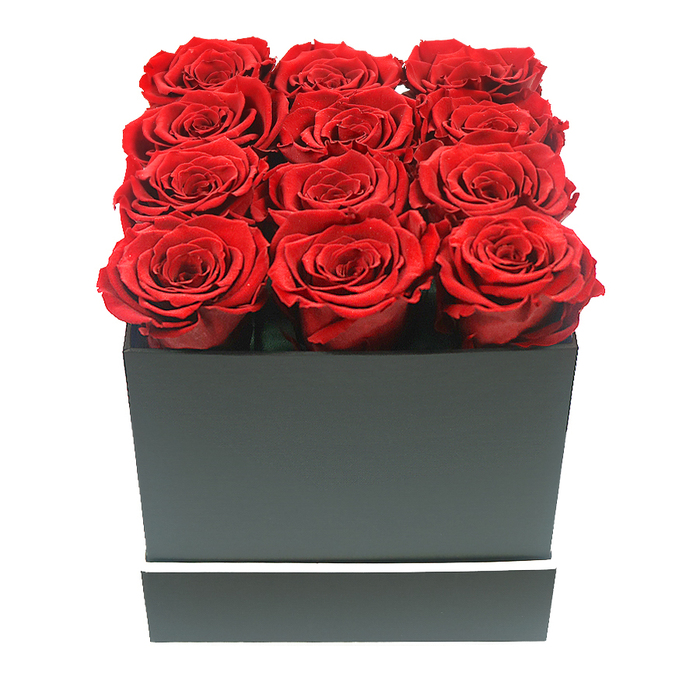 The image presents a carefully composed, eye-level view of a luxurious rose box arrangement that could easily be found on display in a chic Oxford florist just off Cornmarket Street. Twelve extravagant red roses, likely preserved for long-lasting beauty, are meticulously aligned in a matte black square container, filling the entire opening and rising just above the rim in a perfect grid of three by four. Each rose is in full bloom, with densely layered petals unfolding in concentric swirls, their texture soft and velvety like rich velvet drapery in a historic Oxford college hall. The colour is a deep, intense crimson with subtle shifts of tone, from dark wine at the petal bases to brighter red near the edges, giving each flower depth and life. The box itself is minimal and refined, its black surface smooth and untextured, forming clean, geometric lines that contrast beautifully with the organic curves of the blooms. Around the lower section of the box runs a single narrow white band, sharp and even, adding just enough visual detail to elevate the design without distracting from the roses. The arrangement is photographed against a pure white background, with soft, diffused lighting that creates gentle shadows beneath the petals and along the corners of the box, enhancing its three-dimensional form without any harsh reflections. This visual composition feels luxurious and intentional, evoking thoughts of romantic gestures shared along the banks of the Thames in Oxford or celebratory gifts delivered to elegant homes in North Oxford. It suggests enduring love, sophistication, and the thoughtful choice of preserved roses as a long-lasting memento for anniversaries, special birthdays, or Valentine's surprises across the city. Flowers Oxford could easily prepare such a piece for same-day delivery, bringing a touch of modern romance to both historic and contemporary Oxford settings.
