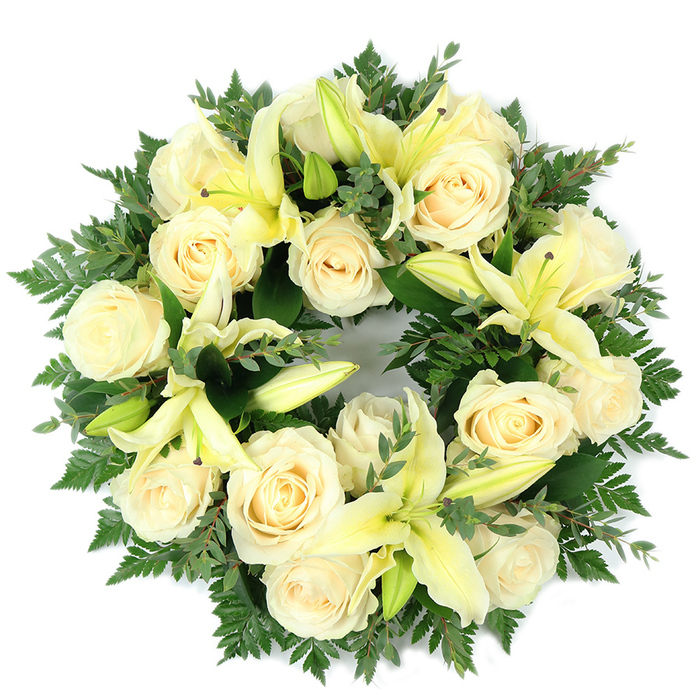 A carefully constructed circular wreath, photographed flat on a white surface and designed as a formal sympathy tribute, combining creamy ivory roses, pale yellow lilies, and a layered, verdant foliage base. Approximately fifteen to twenty full roses form the primary visual weight, their tightly furled petals creating a soft, tactile mass of warm ivory that feels both classic and consoling; interspersed among them are generous trumpet lilies in a delicate lemon hue, some flaring open into elegant arcs while others hold back as buds, contributing subtle architectural variety and promise of continued bloom. The underpinning greens are rich and deliberately varied: delicate fern fronds lend a feathered, airy edge, while thicker, rounder leaves-resembling eucalyptus-supply deeper tones and a cool, silvery-green counterpoint that frames the pale flowers. Shot from above in soft, neutral light, the wreath's dense packing reads as luxurious yet restrained, its perfect circular shape emblematic of continuity and memory. The sensory impression is gentle-the roses suggest a soft, powdery sweetness, the lilies a faint green-citrus lift, and the overall composition conveys serenity rather than exuberance. Crafted with meticulous attention to balance and texture, this wreath is suited to funerals, memorial services, or graveside placements across Oxford, evoking the calm dignity of places like Carfax and the Radcliffe Camera. The design emphasises comfort, purity, and respectful remembrance, offering a graceful and measured visual message for those marking loss.