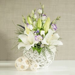An elegant wedding-style centerpiece is captured in soft, natural light, as though set on a polished table in an Oxford townhouse near the High Street. The arrangement is built around striking white lilies, their wide, star-like blooms open and inviting, each petal smooth and satiny with a gentle curve at the tips. Some lilies are still enclosed in elongated, pale green buds, adding height and a sense of unfolding time. Woven among them are lisianthus blossoms in shades of white and cream, several blooms revealing petals tinged with the faintest lavender-purple, like brushed pastel along the edges. Some lisianthus remain in bud form, slender and spiraled, lending a quiet, romantic tension to the composition. Fine sprays of tiny white filler flowers, likely gypsophila, soften the spaces between the larger blooms, while a few deeper purple accents tuck in discreetly, deepening the color palette without overwhelming it. The greenery is abundant yet airy: feathery, fern-like fronds mingle with more structured, rounded leaves, draping gently over the container and giving the piece a natural, garden-inspired flow. All the flowers are gathered in a rounded white vase with an open-weave lattice resembling delicate wickerwork, casting subtle shadows and adding tactile interest. Two pale woven spheres of varying sizes sit beside the vase, their light beige strands twisting into open orbs that echo the vase's pattern. The background is a light beige, linen-textured wall, calming and unobtrusive, letting the whites and greens remain the focus. The arrangement feels tranquil yet celebratory, equally at home on a wedding table in Summertown, a reception by the river in Iffley, or as a refined gift delivered across Oxford.