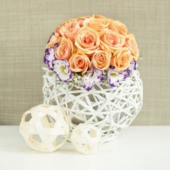 A refined table arrangement rests on a smooth white surface, evoking the quiet elegance of an Oxford wedding breakfast or anniversary dinner tucked away near the Covered Market. At the centre is a carefully sculpted dome of roses in soft peach, apricot, and pale salmon, each bloom pressed closely to its neighbour so that the surface appears almost seamless. Their petals are plush and gently curled, suggesting roses cut at just the right moment, with a fragrance that you can almost imagine drifting across the table. Encircling this rounded crown of roses, a collar of pristine white lisianthus opens outwards, their ruffled petals finely traced with deep violet and purple edges. These bi-coloured blooms introduce a delicate contrast, like ink on parchment, while tiny sprays of white baby's breath weave between them, adding lightness and a touch of whimsy. The flowers rise from an unusual white woven container that swells outwards in a spherical, basket-like form. Made from interlaced strands of wicker or rattan, the structure creates an open lattice that lets the background glow through, giving the impression of air and space beneath the flowers. In the foreground, two decorative woven balls in natural beige hues sit companionably together, one small, one larger, echoing the lattice motif and adding a sculptural element to the composition. The entire arrangement is set against a softly textured beige fabric backdrop, reminiscent of natural linen or burlap, which keeps the focus on the blossoms and their gentle colours. The overall mood is one of calm sophistication and romantic charm, perfectly suited to thoughtful floral styling for events and intimate gatherings across Oxford, from Headington homes to historic venues in the city centre.