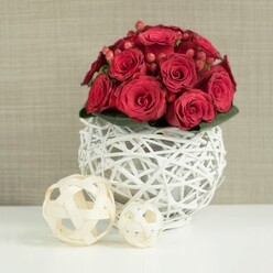 At the heart of the scene is a perfectly rounded centerpiece of red roses, composed with the kind of quiet precision a local Oxford florist brings to wedding tables and anniversary suppers. The roses are a deep, luxurious crimson, their petals thick and velvety, some tightly layered in bud, others gently unfurled to reveal soft shadows at their cores. Threaded between the blooms are fine sprays of red Hypericum berries, each one smooth and glossy, adding a jewel-like sparkle and a slightly different shade of red, as if to echo the changing tones of sunset over Oxford's spires. Just beneath the flowers, a narrow ring of dark green leaves forms a subtle collar, grounding the rich reds and giving the arrangement a sense of completeness. This floral dome is cradled in a distinctive spherical container fashioned from white-painted branches. The twigs are woven into an open lattice sphere, airy and sculptural, allowing glimmers of the stems and foliage to show through and casting delicate shadows onto the pristine white tabletop below. To the left of the vase rest two decorative orbs made from pale, natural wicker, their intertwined strips looser and more rustic, offering a soft, organic contrast to the crisp white sphere. The larger orb sits forward, the smaller one slightly behind, together creating a gentle rhythm of shapes. In the background, a softly textured wall in muted beige or taupe fades into blur, so the eye rests fully on the vivid roses. The image conveys warmth, romance, and carefully considered style, perfectly suited to an intimate wedding breakfast near the Bodleian or an elegant dinner party in an Oxford townhouse.
