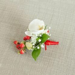 A meticulously designed boutonniere lies centered on a pale beige, woven fabric that looks like finely textured linen, its subtle threads and gentle shadows giving the image a calm, tactile warmth reminiscent of a cosy Oxford townhouse interior. The focal bloom is a pristine white Phalaenopsis orchid, its petals wide, smooth, and almost velvety, opening like a pair of wings around a throat brushed with creamy yellow and soft orange, adding quiet depth at the flower's heart. Surrounding this orchid is a ring of delicate white baby's breath, each tiny blossom forming part of a light, cloud-like cluster that softens the overall silhouette. At one side, a compact cluster of glossy red berries introduces a striking splash of colour, their rounded forms catching the light with a subtle shine that suggests freshness and vitality. Among them, a few pale green berry buds are just beginning to colour, offering a gentle hint of growth and seasonality, perfect for winter weddings or formal events overlooking the spires of central Oxford. Rich green foliage, in varied leaf shapes from broad and pointed to slender and tapered, weaves through the composition, grounding the design in natural abundance. At the base, the exposed stems are neatly wrapped in a smooth red satin ribbon, its surface gleaming softly as it is tied into a small, secure knot, ready to be pinned to a suit lapel. The overall impression is one of refined yet inviting elegance, a small, wearable floral piece that would feel at home at a wedding reception on Banbury Road or a graduation celebration near the University Parks. Crafted with the care you'd expect from an experienced local florist, it balances classic white florals with festive red accents and lush greenery.