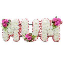 A reverent floral tribute spelling out MUM in large, connected capital letters, lovingly constructed from dense clusters of immaculate white chrysanthemums. Up close, the small chrysanthemum heads form a near-continuous, cloud-like surface across each letter, their petals creating a delicate, textured field that speaks of purity and calm. Each letter's perimeter is softly edged with a ruffled blush-pink ribbon or fabric trim, introducing a comforting warmth against the white blooms, while the dark green structural frame between the letters remains subtly visible to support the standing tribute. Three coordinated accent bouquets interrupt the white with bright, emotional colour: a compact spray of deep fuchsia-pink roses, fern-like greenery and glossy reddish-orange hypericum berries sits at the upper left of the first 'M'; the same combination nestles into the base curve of the 'U'; and a final mirrored cluster decorates the upper right of the second 'M'. The roses' velvety petals, the crisp fern fronds, and the shiny berries give pleasing tactile variety beneath gentle indoor lighting you might find in a chapel near Summertown or a family home around Iffley. This is the kind of arrangement families from across Oxford - from Jericho to Headington - request when they want a personalised, heartfelt floral message for a funeral or memorial. Prepared with the care you'd expect from a local florist such as Flowers Oxford, the piece balances classic sympathy symbolism with subtle colour to honour a beloved mother with dignity and affection.