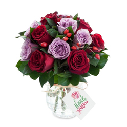 A richly coloured bouquet crafted in a rounded, compact style: velvety red and burgundy roses are paired with gentle lavender and mauve roses, their petals unfurling in layered spirals that catch the light with a satin sheen. Interlaced between the blossoms, deep green foliage - likely Ruscus or Salal - creates contrasting planes of cool, glossy texture that support and lift the flowers. Scattered through the mass of roses are clusters of small, vivid red-orange rose hips, their firm, glossy skins introducing a playful, rustic note that offsets the arrangement's elegance. The stems are gathered into a reflective mercury-glass vase, its mottled, antique-silver surface shimmering subtly; the vase's rounded base and slightly narrowed neck are wrapped with natural twine or raffia, from which a white rectangular tag hangs, annotated with a tiny stylised rose sketch and a blank space for a message. Photographed against a spotless white backdrop, the colours feel both saturated and true - a composition that reads as romantic, considered, and premium. As a local florist would note, the bouquet would travel well across Oxford - perhaps to a sunlit shop on Cornmarket Street or delivered to a riverside flat near Port Meadow - its compact shape and sturdy vase making it easy to place on a mantel or dining table. The overall impression is one of warm affection and refined taste: a bouquet to mark birthdays, anniversaries, or quiet moments when you want to say, simply and beautifully, you are thought of.