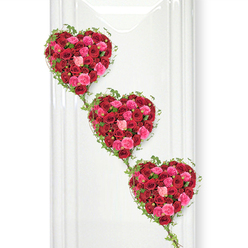In this refined funeral tribute, three heart-shaped floral designs are arranged in a gentle downward sweep, from the upper left to the lower right, like a quiet procession of love. Each heart is completely filled with roses in tones of rich, velvety crimson and lively pinks, the petals layered tightly together until the surface looks like a plush, living fabric. Some roses curl softly into half-open buds, while others are fully unfurled, revealing spiralled centres and delicate edges that catch the light. Around every heart, slim stems of green ivy form a delicate border, their small, glossy leaves adding a natural frame and a hint of wildness to the otherwise precise shape. These ivy tendrils stretch down between the hearts, binding them together in a graceful chain that can evoke generations of family, or the unbroken connection between the living and the departed. The background is a perfectly smooth, bright white plane, reminiscent of a freshly painted wall in an Oxford funeral chapel or a serene space near the Botanic Garden, allowing the reds, pinks, and greens to appear especially vivid and pure. Soft, even lighting falls across the flowers, emphasizing the depth of colour and the subtle shadows between petals. The mood is tender yet composed, ideal for honouring a loved one with a tribute that speaks clearly of affection, loyalty, and remembrance. Created with the eye of a skilled Oxford florist, this triple-heart arrangement brings a sense of dignity and warmth to any memorial or celebration of life.