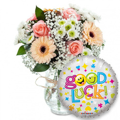 Soft morning light seems to settle over this comforting 'Good Luck' gift: a lush bouquet positioned slightly left of centre in a clear glass vase, its neck finished with rustic twine, the stems visible and neatly gathered below. As your florist I note the palette - pale peach and powder pink roses with tightly furled petals, paired with peachy-orange Gerbera daisies whose dark brown centres offer a warm focal point. Sprays of white Gypsophila tumble through the arrangement like airy lace, while compact clusters of white and lime-green chrysanthemum-like blooms add bright punctuation and crisp texture. Deep green foliage threads through the flowers, framing each bloom and lending a fresh, garden-cut scent that complements the roses' gentle perfume.To the right and overlapping the lower edge of the bouquet a round silver Mylar balloon gleams, its metallic face peppered with cheerful motifs: tiny hearts in blue and pink, stars in yellow and blue, and small multicoloured abstract shapes. Written across it in a playful, multicoloured typeface is "Good Luck!" - the letters bounce in green, orange, red and blue, and yellow smiley faces are playfully used as letterforms, lending friendly personality. The KALEIDOSCOPE brand mark is visible at the bottom rim of the balloon.This composition radiates encouragement and quiet celebration - perfect for sending to students before exams, colleagues starting new roles, or anyone facing a milestone. As Flowers Oxford often arranges for deliveries around Jericho, Summertown and the University Parks, the bouquet reads as both intimate and uplifting - a tactile, fragrant message of support made for neighbourhood moments.