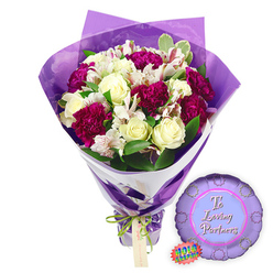 Set against a pure white background, this image shows a romantic floral gift set that feels perfectly at home in Oxford's blend of tradition and quiet romance. The hand-tied bouquet is full and carefully sculpted, built around creamy white roses whose tightly curled petals spiral inward with an air of classic elegance. Nestled between them are generous clusters of deep magenta and burgundy carnations, their frilled, densely layered petals adding bold colour and a velvety texture that suggests warmth and commitment. Threaded throughout are white alstroemeria, or Peruvian lilies, each bloom marked with fine reddish-brown streaks and delicate freckles on its inner petals, offering a refined, almost painterly contrast that a local florist near the Covered Market might take pride in. Lush green foliage, some leaves solid and others softly variegated at the edges, surrounds the blossoms, framing them in fresh colour like greenery along the towpath by the Thames. The flowers are wrapped in two distinct layers: an inner sleeve of smooth, clean white paper that cradles the stems, and an outer layer of translucent, slightly crinkled purple cellophane that rises around the arrangement, catching the light and giving the bouquet a subtle glow. At the base, a bright green satin ribbon is tied neatly around the stems, finished with a small, precise bow from which hangs a light brown rectangular gift tag, waiting for a personal note. To the right, a round metallic purple foil balloon with a scalloped, medal-like edge rests lightly on the surface. In its centre, a softer lilac circle holds the message "To Loving Partners" in elegant pink script, completing a thoughtful romantic ensemble ideal for anniversaries, proposals, or celebrations delivered across Oxford's neighbourhoods, from Summertown to Cowley Road.