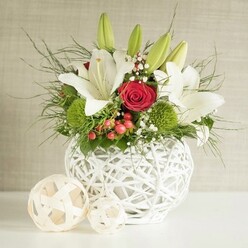 On a bright, uncluttered surface, a beautifully composed floral centerpiece rests in a distinctive round white vase, its open-weave structure reminiscent of delicately woven branches, much like the hand-crafted pieces often seen in Oxford's Covered Market. The vase is filled to overflowing with a harmonious mix of white, red, and green, arranged with the quiet confidence of an experienced wedding florist. At the heart of the display, several white lilies rise gracefully, some in full, fragrant bloom with slightly speckled petals and golden stamens, others still in elongated green bud form, adding height and a sense of ongoing life to the design. Tucked between the lilies are velvety red roses, with one central bloom drawing the eye, its deeply layered crimson petals evoking warmth, devotion, and the intimacy of a small Oxford wedding breakfast near Christ Church Meadow. Around these focal flowers, bright green pompon-like blossoms-likely green dianthus or similar chrysanthemums-offer a playful, rounded texture that contrasts with the lilies' wide, elegant petals. Airy sprigs of white baby's breath float throughout the bouquet like tiny stars, softening the rich colours, while clusters of glossy red berries introduce a subtle seasonal touch, ideal for winter receptions or festive table settings. Lush greenery in various shapes spills from the arrangement: fine, feathery fronds, arching stems, and broader deep green leaves create volume, structure, and a natural garden feel. The white woven vase rests beside two smaller decorative spheres made from pale, intertwined strands, echoing its texture. Against a gentle neutral background, the entire centerpiece feels calm yet celebratory, perfectly suited to wedding tables and special gatherings across Oxford, from Jericho townhouses to riverside venues near Iffley.