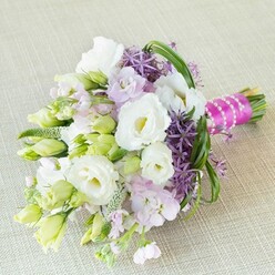 Against a softly woven, neutral-toned backdrop that calls to mind the charm of Oxford's historic interiors, a radiant bouquet is captured with natural grace. Creamy white lisianthus flowers bloom wide, their petals gently ruffled as if shaped by a breeze from University Parks. Surrounding them, clusters of unopened lisianthus buds in pale green and white add promise and subtle fragrance, weaving together past and future. Fluttering amidst these central blooms are soft lavender-pink stocks, their airy petals lending a dreamy feel akin to oxford wildflower meadows in spring. Striking deep purple allium flowers stand boldly, each tiny star-shaped floret adding a geometric structure and vivid contrast-an architectural element reminiscent of the city's famed spires. Fresh lime-green buds and textured greenery peek out in various stages, echoing the renewal of local botanic gardens. The bouquet's arching blades of slender, glossy bear grass loop with quiet energy, unifying the palette. At the base, a bright fuchsia satin ribbon wraps the stems tightly, interlaced with a luminous string of white pearls that shimmer with understated elegance. This bouquet, crafted by a passionate Oxford florist, evokes themes of romance, unity, and celebration. Ideal for a local wedding, bridal shower, or special event, it offers not just beauty but a sense of place-perfect for delivery throughout Oxford's storied neighborhoods and colleges.