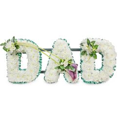 This image shows a carefully crafted funeral flower tribute that spells DAD in bold, upright letters, resting against a bright white background so every detail stands out. Each letter is densely filled with immaculate white chrysanthemums, or similar small button flowers, their tightly layered petals forming a plush, almost cushion-like surface that suggests softness and care. Encircling the outside of every letter is a vibrant deep green ribbon, neatly pleated to give a ruffled edge, which adds structure, contrast, and a quiet echo of the greenery found in Oxford's parks and riverside walks. Behind the flowers, a dark supporting frame and horizontal bars can just be made out, indicating that the piece is mounted on a stand, as it might be when placed beside a coffin or at a graveside service. The top left of the first D is adorned with a dainty cluster of white freesias and fresh foliage, their slender buds and open blooms resting lightly on the white chrysanthemum base. Two sweeping green stems arc from this first letter across towards the central A, where they form a graceful diagonal line that draws the eye. At their meeting point sits an expressive spray of flowers: two rich, deep purple calla lilies with sculpted, trumpet-like heads, tucked among more white blossoms and glossy leaves, offering a touch of solemn elegance and colour. On the upper right of the final D, another cluster of white blooms and greenery mirrors the first, creating a sense of balance. The tribute feels calm, respectful, and deeply affectionate, the kind of personalised farewell a family in Oxford might choose to remember a devoted dad with quiet pride.