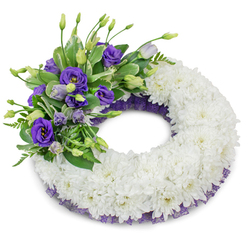 Set against a stark white backdrop, this circular sympathy wreath is a study in restraint and thoughtful detail, hand-assembled to convey calm and remembrance. The principal body forms a continuous ring of pristine white cushion chrysanthemums - each tight, ruffled bloom layered to produce a dense, velvety field that evokes purity and lasting affection. Anchoring the upper-left quadrant is a rich spray of purple Eustoma (Lisianthus), displaying a gentle gradient from deep violet blooms with soft yellow centres to pale lavender buds that suggest quiet hope. The purple cluster is lightly threaded with mixed greenery - variegated foliage and feathery fern-like leaves - bringing botanical texture and a subtle, fresh scent that calls to mind early walks through University Parks. A delicate border of crinkled dark-purple mesh or lace peeks along the inner and outer edges, complementing the lisianthus and adding a refined frame to the white base. Soft studio lighting casts a gentle shadow beneath the wreath, highlighting its three-dimensional structure and the meticulous hand-placed flowers. As a local florist in Oxford I often prepare pieces like this for services at the University Church or for intimate tributes near Jericho and the Covered Market; Flowers Oxford carefully balances colour, symbolism and proportion so the wreath reads as both elegant and consoling. It is intended to express heartfelt sympathy and enduring remembrance with dignity and grace.