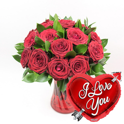 A luxurious romantic arrangement features a clear, fluted ruby-red glass vase brimming with deep red roses, gathered so closely together that they form a rich, velvety dome of colour. Each rose varies slightly in its stage of bloom: some are open and full, with satiny petals sweeping outwards, while others remain gently cupped, holding their fragrance and promise a little closer. Between the blooms, dark emerald foliage arches and curves, giving the design texture and contrast, much like the leafy avenues around University Parks in Oxford. The red glass vase itself has soft vertical ridges that catch and bend the light, lending a sense of movement and depth to the base of the display and subtly echoing the stems nestled within. Against a crisp white background, every shade of red and green appears especially vivid, as if the bouquet is illuminated by fresh morning light. In the lower right foreground, overlapping the line of the vase, a shiny, metallic red heart-shaped balloon leans in toward the flowers, as though drawn by their warmth. On its reflective surface, the words "I Love You" are written in flowing white script, easy to read and full of feeling. A stylised silver Cupid's arrow runs diagonally through the heart, symbolising love that has struck true. The image evokes the moment just before a surprise delivery arrives at a front door in Summertown or Headington: a classic pairing of roses and heart motif, ideal for marking anniversaries, Valentine's Day, or any occasion where love needs to be seen and felt immediately.