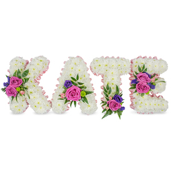 A lovingly crafted standing tribute that spells KATE, each letter sculpted from thousands of snow-white chrysanthemums to form a plush, three-dimensional surface against a bright white backdrop. A whisper of pale pink satin ribbon outlines the letters' edges, softening their silhouettes and imparting a gentle rosé halo around the crisp white blooms. Contrasting clusters punctuate each letter: rich fuchsia-pink roses, tender pastel spray roses, and fine deep purple or indigo sprays-likely statice or small lisianthus-nestled into pockets of greenery. Textural foliage, including delicate fern fronds and rounded eucalyptus leaves, frames the clusters and adds depth and a faint, calming eucalyptus scent that blends with the rose fragrance. Positioning is deliberate and balanced: the diagonal stroke of the K carries a bold cluster, the A is accented near its lower right curve, the T's base is grounded by a bloom group, while the E features two arrangements on its middle and bottom bars, lending visual cadence across the word. The lighting is soft and directional, making the white chrysanthemums glow while allowing the pinks and purples to read with comforting clarity. Prepared with the attention to ceremony one expects from a long-standing Oxford florist, this tribute would be suitable for a chapel service near the Covered Market or a memorial at St Giles-Flowers Oxford can recreate this design for local services, ensuring the composition reads respectfully from the front pew or graveside.