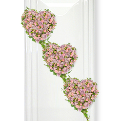 A refined sympathy arrangement features three interlinked hearts of roses, suspended visually against a crisp white backdrop that highlights their soft colours and careful craftsmanship. Each heart is a full, plush cushion of miniature roses in muted lavender, dusty mauve, and gentle blush pink, the petals layered densely to form a velvety surface that feels both comforting and luxurious. The hearts descend diagonally from top left to bottom right, giving the tribute a cascading effect, as if love were gently flowing from one heart to the next. Around and between them, slender strands of fresh green ivy weave in looping curves, defining the edges and creating natural bridges, the glossy foliage adding depth and a touch of woodland calm, like a walk through Christ Church Meadow in Oxford on a quiet day. Subtle reflections in the bright background suggest a polished wall or glass panel, adding a contemporary note without distracting from the flowers themselves. Every bloom looks carefully placed, with no gaps, so that the hearts appear solid yet soft, a visual expression of closeness and unity. This triple-heart floral tribute would sit beautifully beside a coffin or at the front of a chapel in Oxford, offering a serene message of enduring devotion, shared memories, and family ties. The gentle pastel tones and flowing greenery evoke peace and tenderness, making it especially fitting for commemorating a life well lived with grace, love, and gratitude.