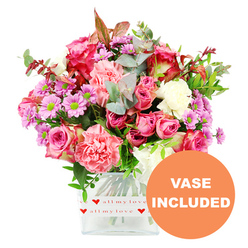 A vibrant mixed bouquet fills a clear, square glass vase, photographed against a spotless white backdrop so every detail stands out, as if displayed in a light-filled Oxford florist's studio. At its heart are generous pink roses, some a rich magenta with darker-tipped petals, others a softer, romantic blush that feels like a gentle embrace. These are nestled among plush, ruffled carnations in coral, peach-pink, and soft rose tones, their layered petals giving the arrangement a luxurious fullness. Dancing between the larger blooms are clusters of daisy-like chrysanthemums in shades of lavender and pale purple, each framed by luminous green centres that bring a lively sparkle, while creamy white flowers offer calm moments of light throughout the bouquet. The flowers are supported by abundant greenery: rounded eucalyptus leaves with their cool, silvery-green sheen, deeper green foliage adding depth, and a thread of reddish-brown leaves that hints at cosy autumn evenings by the colleges. The bouquet is presented in a transparent, rectilinear vase whose base is wrapped in a neat white band patterned with bright red hearts and the affectionate script "all my love," making the intention of the gift unmistakable. In the bottom right corner of the image, an orange circular overlay clearly reads "VASE INCLUDED" in bold white text, reassuring anyone ordering same-day flower delivery in Oxford that this is a complete, ready-to-display present for birthdays, anniversaries, or simply brightening a home near Summertown, Headington, or along the Cowley Road. The overall effect is one of thoughtfulness, colour, and tender emotion.