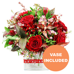 Against a pure white background, this romantic bouquet feels like a heartfelt love letter expressed in flowers, the kind of design a local Oxford florist might prepare for a proposal by the Radcliffe Camera or an anniversary dinner on Cowley Road. At the centre are sumptuous red roses, some tightly furled, others beginning to open into layered rosettes, their velvety petals creating soft shadows and depth. Nestled among them are smaller deep red blooms resembling alstroemeria or similar multi-petalled flowers, each one adding a sculptural quality and enhancing the passionate red palette. Threaded between these larger blooms are delicate sprays of tiny pale pink and white waxflower-style blossoms, their starry heads and slender stems lending a light, almost lacy texture. Around the flowers, an abundance of green foliage forms a generous, rounded dome: glossy leaves, finer sprigs, and arching stems all working together to frame the roses. Striking burgundy and reddish-brown leaves with gentle variegation peek through the greenery, introducing depth and a refined, dramatic edge that feels almost like the contrast of old stone and fresh greenery in Oxford's college gardens. The bouquet is anchored in a simple, contemporary clear glass vase with a rectangular base, allowing neatly cut stems to be seen and lending a polished, modern finish. A festive red ribbon printed with small white heart outlines wraps around the vase, adding a playful, clearly romantic touch. In the lower right corner, an orange circular label with bold white uppercase text clearly states "VASE INCLUDED," signalling that this arrangement arrives beautifully presented and ready to display, perfect for same-day romantic flower delivery across Oxford.