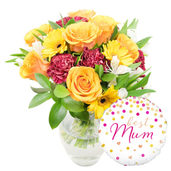 Against a pure white studio background, a lush arrangement leans gently to the centre-left with a glossy round balloon playfully positioned at the lower right, overlapping the vase's base. The flowers read as an intimate, sun-warmed composition: large peachy-orange roses with velvet-soft petals form the heart of the bouquet, their layered spirals suggesting calm and celebration; golden-yellow gerberas peek through like small suns, their dark centres giving crisp graphic contrast; and deep maroon carnations with densely ruffled edges bring a plush, tactile richness. Interwoven among these principal blooms are sprays of white alstroemeria, their narrow petals flecked with subtle markings, which lift the palette with delicate lightness. Surrounding the blooms, abundant green foliage-glossy broad leaves and fine sprigs-creates a verdant frame and a sense of freshly cut stems, visible through a simple clear glass vase filled with water. The vase's transparency highlights the clean, trimmed stems and the care of the arranging process. On the white balloon, jaunty gold and hot-pink polka dots and tiny hearts scatter like confetti; the phrase "best Mum" is scripted in flowing hot-pink lettering that reads as both affectionate and celebratory. This is the sort of bouquet that feels right for a mother's birthday or Mother's Day delivery across north and east Oxford - from Summertown's terraces to the friendly streets around Cowley - offering bright colour, scent, and an unmistakable message of love.