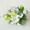 On a soft, off-white linen surface, a horizontally arranged bouquet lies like a small, cultivated patch of meadow. The dominant white Oriental lilies, some fully open with their broad, curling petals and bright orange-yellow stamens, show tiny dark pollen flecks and delicate maroon speckles toward their throats. Between these luminous lilies, white lisianthus edged in violet and deeper purple provide a painterly counterpoint-each ruffled petal moving from a creamy centre to a wash of lavender at the margins. Several lisianthus buds remain tightly furled, promising later blooms and a sense of time within the bouquet's composition. Tiny clusters of Gypsophila (baby's breath) dot the arrangement, lending an ethereal, cloud-like texture that lifts the heavier blooms.

The foliage is varied and deliberately textural: glossy, broad leaves (like salal or ruscus) anchor the shape; slender, feathery foliage and spike-like grasses give direction and movement. The stems are neatly wrapped in a white satin ribbon, completed with small, gem-like pin details that twinkle gently in soft light. The tactile contrasts-silky lily petals, ruffled lisianthus edges, airy gypsophila and crisp greenery-create a bouquet that feels fresh and intentional. It suggests a quiet Oxford morning, as if gathered from a market stall off the High Street and arranged with considered care, perfect for weddings, anniversaries or a thoughtful, elegant gift.
